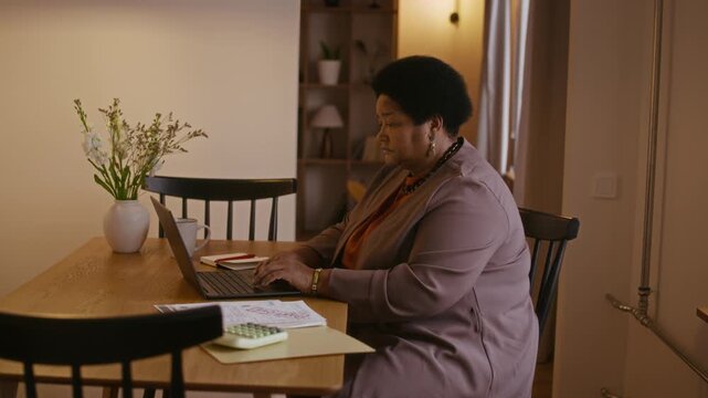 Side view medium slowmo shot of confident senior African American woman sitting at desk in home study, typing on laptop and reviewing financial documents while planning family budget