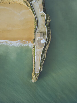 Aerial view of a striking contrast between the golden beach, the rugged cliffs, and the azure sea surrounding the Nossa Senhora da Rocha chapel, Porches, Faro, Portugal.