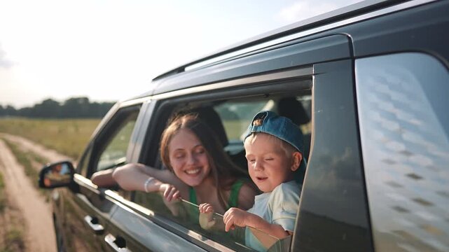 Mother and son look from car window during road trip. Happy family travels through countryside. Boy with cap enjoys journey. Summer vacation on rural road. Family shares happy car trip.