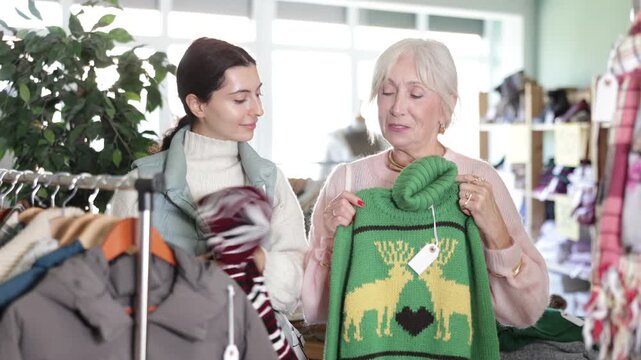 Woman and an elderly customer look at sweaters in a store, discussing the style and quality of knitwear. Customers choose warm knitted sweaters with different patterns. High quality 4k footage