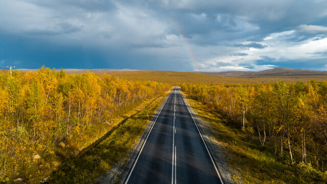 Aerial view of road cutting through the yellow autumn forest with a faint rainbow in the background, Enontekio Kilpisjarvi, Lappi, Finland.