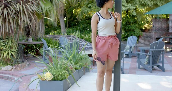 Adult African American woman noticing camera walking on patio in shorts with shades posing at post