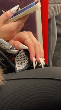 closeup manicured hands scrolling phone on red seat inside metro carriage, long white acrylic nails with rings, winter coat sleeve, casual commuter vibe, Video taken on mobile device.