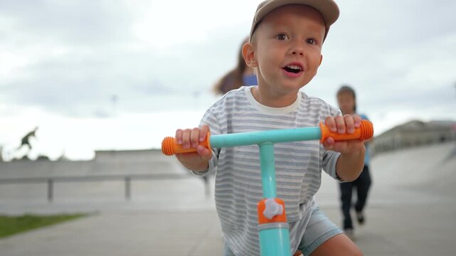 Boy rides balance bike at skate park while mother and sister watch. Child learns riding with family support. Toddler practices bike on ramp. Mother watches boy learning balance bike outdoors.
