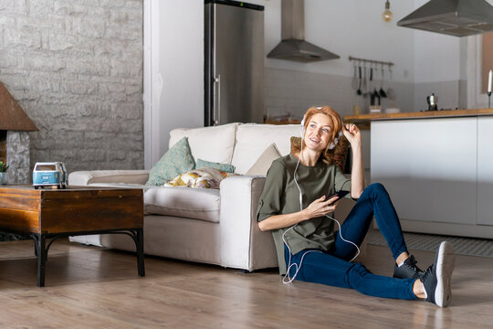 Young woman sitting on floor at home, using smartphone, wearing headphones