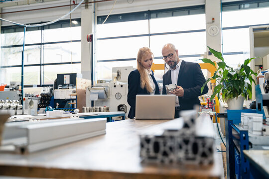 Businessman and businesswoman with laptop examining workpiece in factory