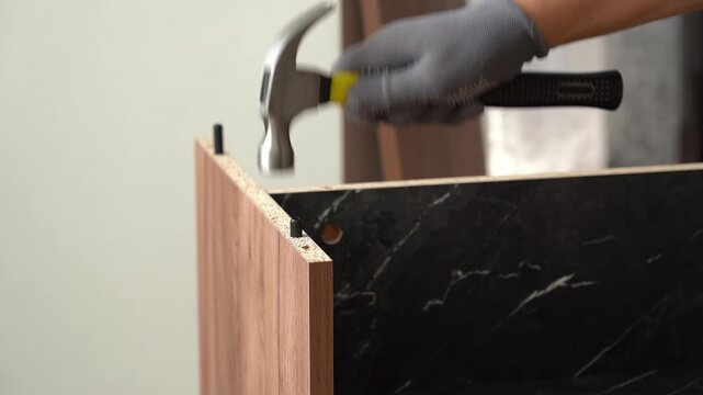 Male hands assemble wooden nightstand using hammer at home. Installing furniture dowel pin. Process of assembling furniture