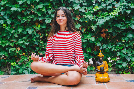 Smiling girl meditating by Buddha sculpture in backyard