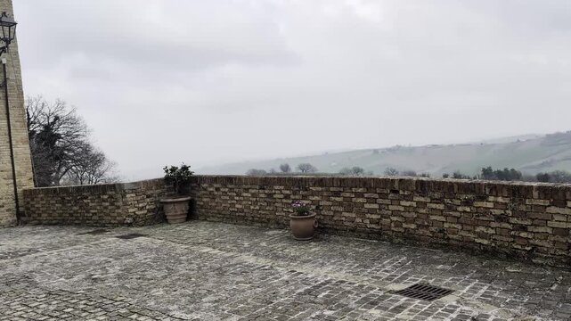 Una terrazza in pietra dell'antico borgo medievale, affacciata su un paesaggio collinare immerso nella nebbia mattutina. 