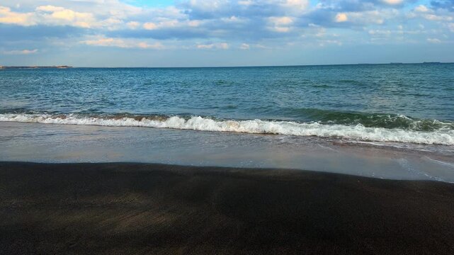 Black sand beach under cloudy sky with distant ships. Wide shot of coastline with dark volcanic sand, breaking waves, and industrial ships on the horizon.