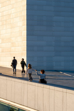 People and a pedestrian move along a minimal urban walkway as a shadow falls across a modern wall in bright sunlight beside clean architecture