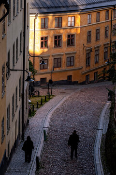 People walking as a pedestrian crosses a Stockholm street of cobblestone in a historic alley at evening where urban architecture shapes old town mood