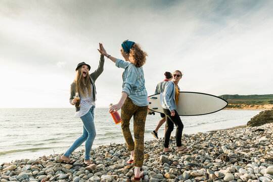 Excited friends with surfboard and drinks on stony beach