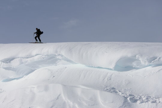 Aerial view of a lone skier cresting a snow-covered ridge under a vast sky, a study in contrasts of dark figure against the bright, undulating white, Echenevex, Auvergne-Rhone-Alpes, France.