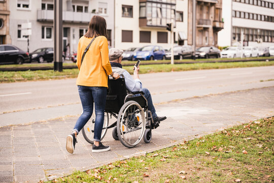 Back view of young woman pushing senior man with smartphone in wheelchair on pavement