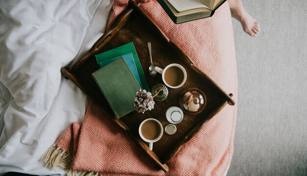 Wooden tray holds coffee cups, books, and flowers on a cozy bed. Soft blankets and neutral tones create a calm, relaxing atmosphere. The scene suggests quiet morning reading or peaceful downtime