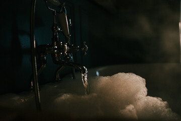 Dark, moody bathtub with foamy water and dripping faucet. Warm glow highlights bubbles and vintage brass fixture. Empty scene evokes relaxation or solitude in dim lighting. Perfect for spa, wellness © Reuben
