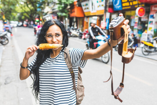 Vietnam, Hanoi, young woman taking a selfie with old-fashioned camera on the street eating a corn cob