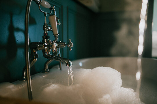 Vintage bathtub fills with soapy water, steam rising gently. Elegant brass fixtures gleam under soft, warm lighting. The scene evokes relaxation and comfort. Ideal for spa, wellness