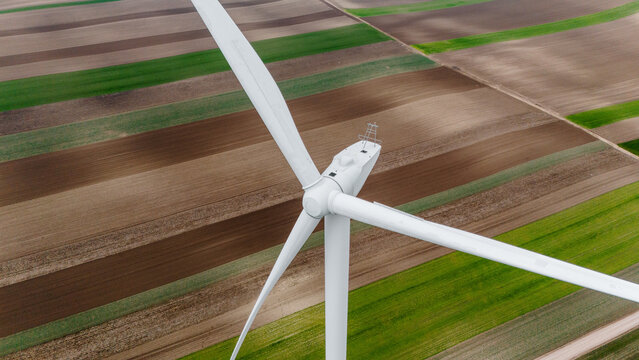 Aerial view of a colossal wind turbine standing tall amidst the patchwork quilt of agricultural fields, its white blades contrasting with the earthy tones, Alibunar, Vojvodina, Serbia.
