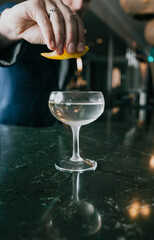Obraz premium Hand squeezes lemon over cocktail glass. Liquid drips into clear drink. Dark bar background with warm lights. Bartender prepares refreshing beverage. Perfect for restaurant, bar