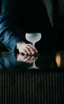 Hand grips frosty cocktail glass on dark reflective bar surface. Dark ambiance with moody lighting highlights bartender's action. Glass reflects in polished surface below. Scene evokes sophistication