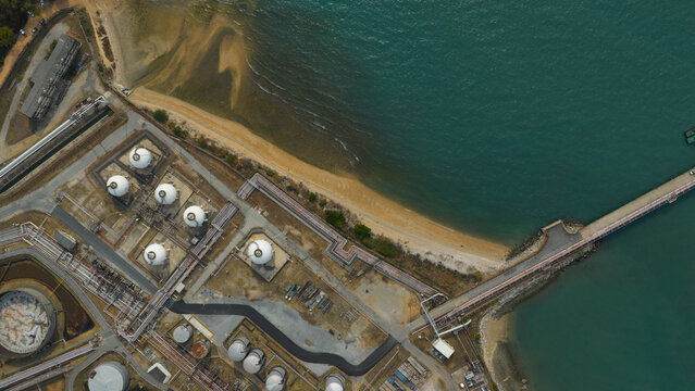 Aerial view of oil storage tanks contrasting with the turquoise sea and sandy shore, Pengerang, Malaysia.