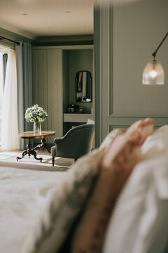 Soft focus on cozy bedroom with muted green walls and vintage chair. White bed in foreground creates inviting atmosphere. Floral arrangement adds delicate charm near window