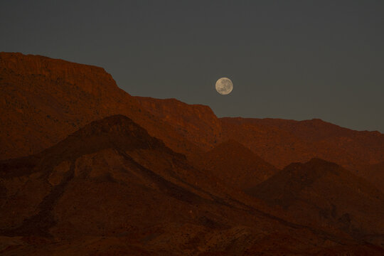View of the full moon glows against the red mountains, casting long shadows across the rugged terrain in Brandberg, Erongo Region, Namibia.