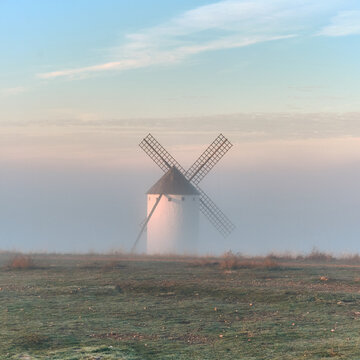 View of a solitary white windmill shrouded in ethereal mist, its sails reaching towards the pastel sky, a timeless sentinel in the tranquil landscape, Campo de Criptana, Castile-La Mancha, Spain.