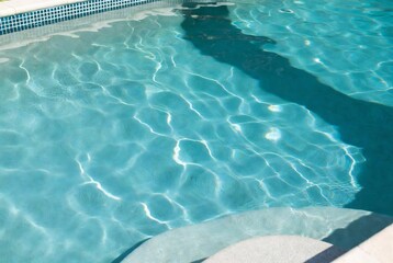 A swimming pool with sunlight reflecting on the water. Someone is in the pool, creating ripples and shadows. It&rsquo;s a sunny day at a swimming pool.