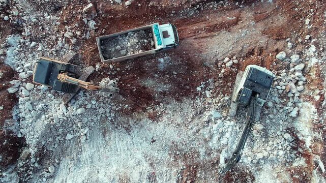 Top-down drone view of heavy construction machinery operating in quarry or excavation site. Two excavators are digging and loading rocks into dump truck surrounded by piles of stone and exposed earth