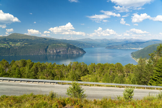 View of a winding road cuts through verdant hillsides, offering a glimpse of the tranquil Innvik Fjord waters reflecting the azure sky, Utvik, Vestland, Norway.