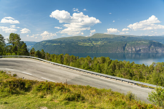 View of a winding road curves through lush green hills, leading towards a serene lake of Innvik Fjord reflecting the distant mountains, Utvik, Vestland, Norway.