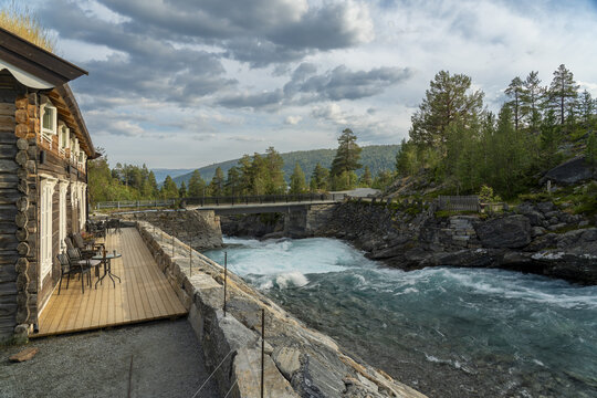 View of a rustic log cabin with a grass roof overlooks a powerful turquoise Tora river rushing under a stone bridge, Billingen, Norway.