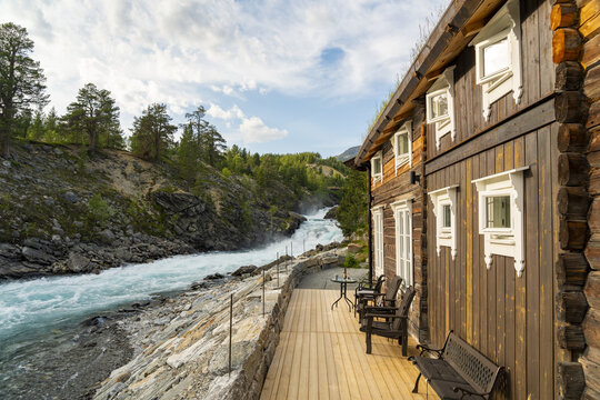 View of a rushing Tora river framed by a rustic wooden cabin with white trim and a backdrop of lush green forest, cascading down rocky slopes, Billingen, Norway.