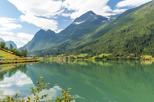 View of serene turquoise glacial lake waters mirroring the towering, forest-clad mountains under a sky brushed with clouds, a picturesque tableau, Floen, Olden, Norway.