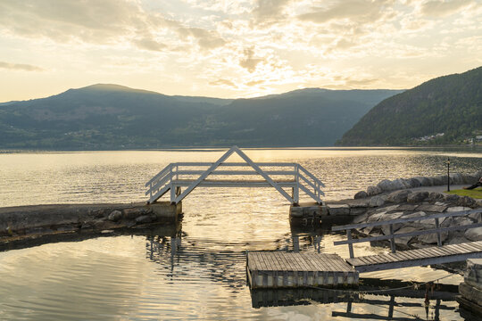 View of a calm lake reflecting the soft golden light of the setting sun, framed by rugged mountains and a quaint white bridge, Innvik Fjord, Vestland, Norway.