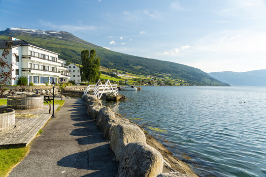 View of serene waters reflecting the sky's azure hues, framed by lush green mountains and charming buildings, Innvik Fjord, Vestland, Norway.