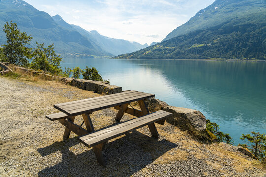 View of a picnic table perched above the shimmering, turquoise waters of the fjord, framed by towering mountains and verdant forests, Innvik Fjord, Vestland, Norway.
