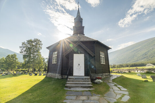 View of the dark, wooden Nordberg Church stands proudly under a bright sun, its white door a stark contrast against the lush green landscape, Nordberg, Innlandet, Norway.