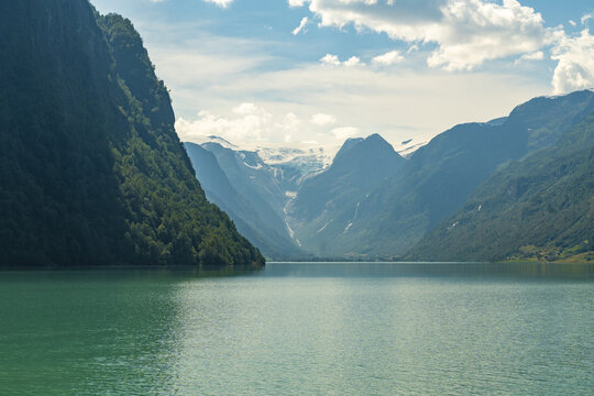 View of tranquil turquoise waters reflecting the towering green mountains against a backdrop of snow-capped peaks and a sky streaked with clouds, Oldevatnet, Vestland, Norway.