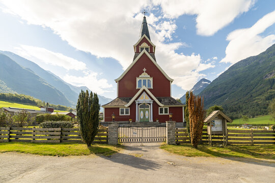 View of a striking red Olden Old Church with a tall spire stands proudly against a backdrop of majestic green mountains and blue skies, Olden, Vestland, Norway.