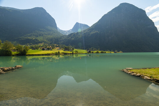 View of turquoise waters mirror the towering mountains under a bright sun, nestled among green fields and trees, Oldevatnet, Vestland, Norway.