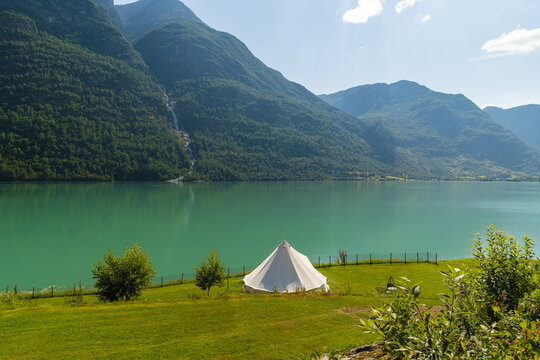 View of emerald lake mirroring majestic mountains, a white tent pitched on lush green shores under a serene sky, Oldevatnet, Vestland, Norway.