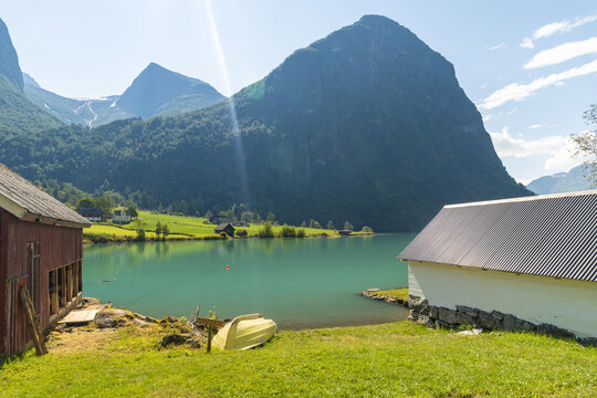 View of turquoise waters reflecting the majestic mountains under a sunny sky, with rustic boathouses dotting the grassy shore, Oldevatnet, Vestland, Norway.