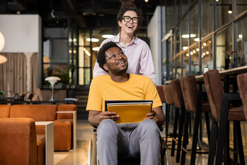 Woman helping disabled coworker by pushing his wheelchair in coworking interior