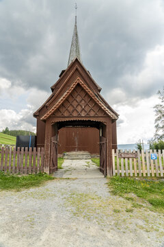 View of an old wooden Hegge Stave church with a tall spire rises against the cloudy sky, framed by a rustic wooden fence and gate, Hegge, Innlandet, Norway.