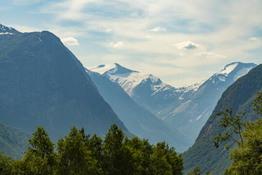 View of towering, verdant mountains rise dramatically, some capped with pristine snow, creating a serene vista in Hjelldalen, Vestland, Norway.