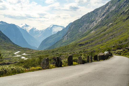 View of a winding asphalt road lined with monoliths through a lush valley leading to snow-capped mountains under a blue sky, Hjelldalen, Vestland, Norway.